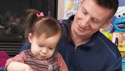 Child with hearing aid sitting on man's lap reading a book